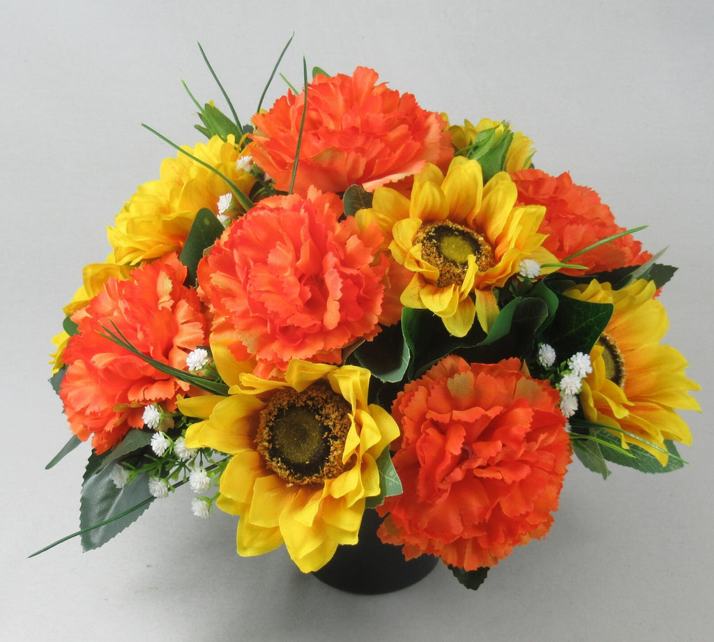 Artificial grave arrangement in dark pot with orange carnations, yellow sunflowers, and white accents