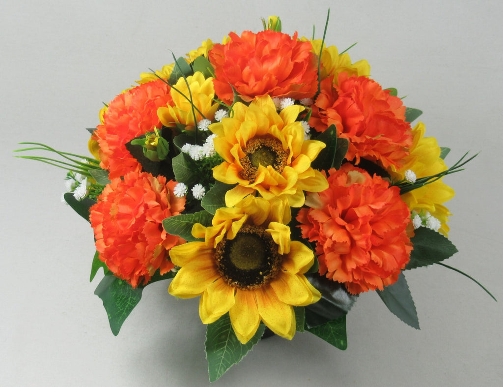 Artificial grave arrangement in dark pot with orange carnations, yellow sunflowers, and green leaves