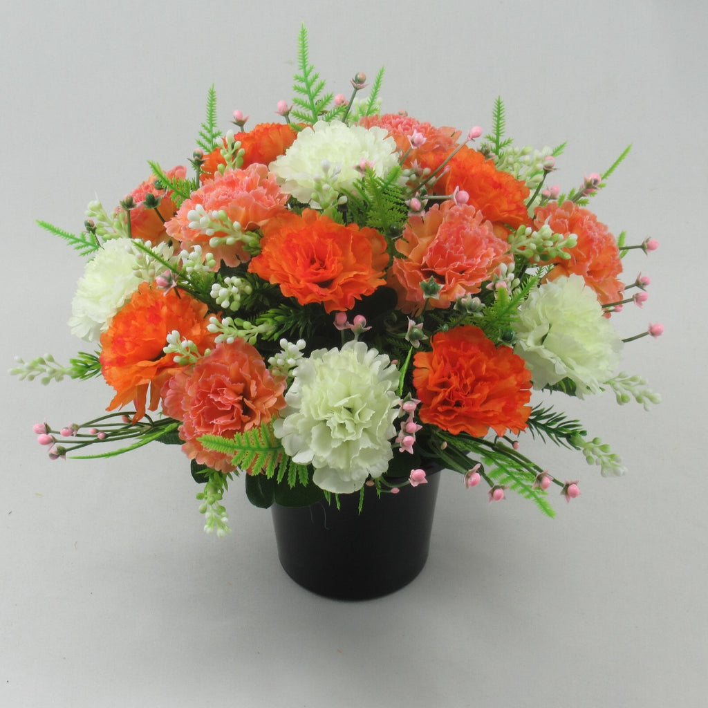 Artificial silk flowers in black grave pot, orange and white blossoms with green foliage