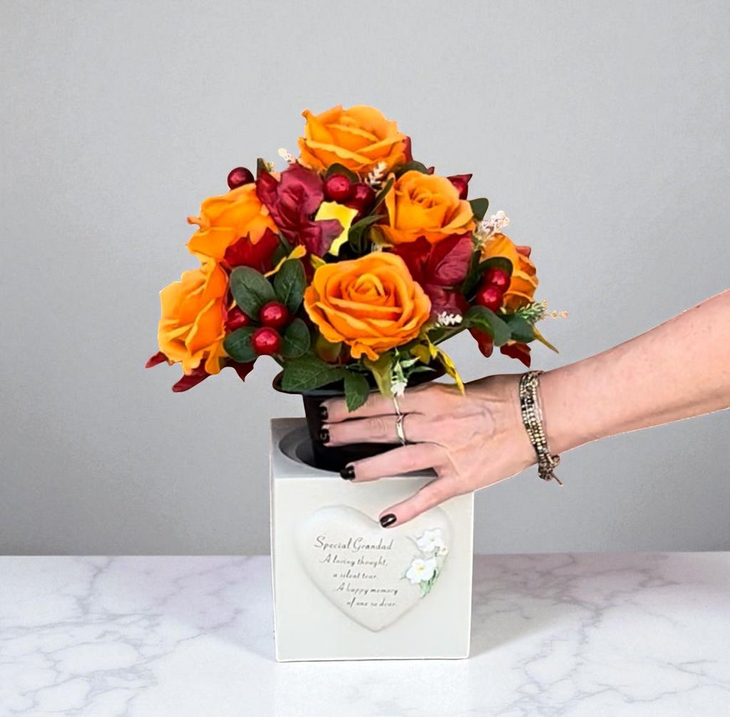 Hand placing orange artificial roses with red berries into a white memorial grave pot