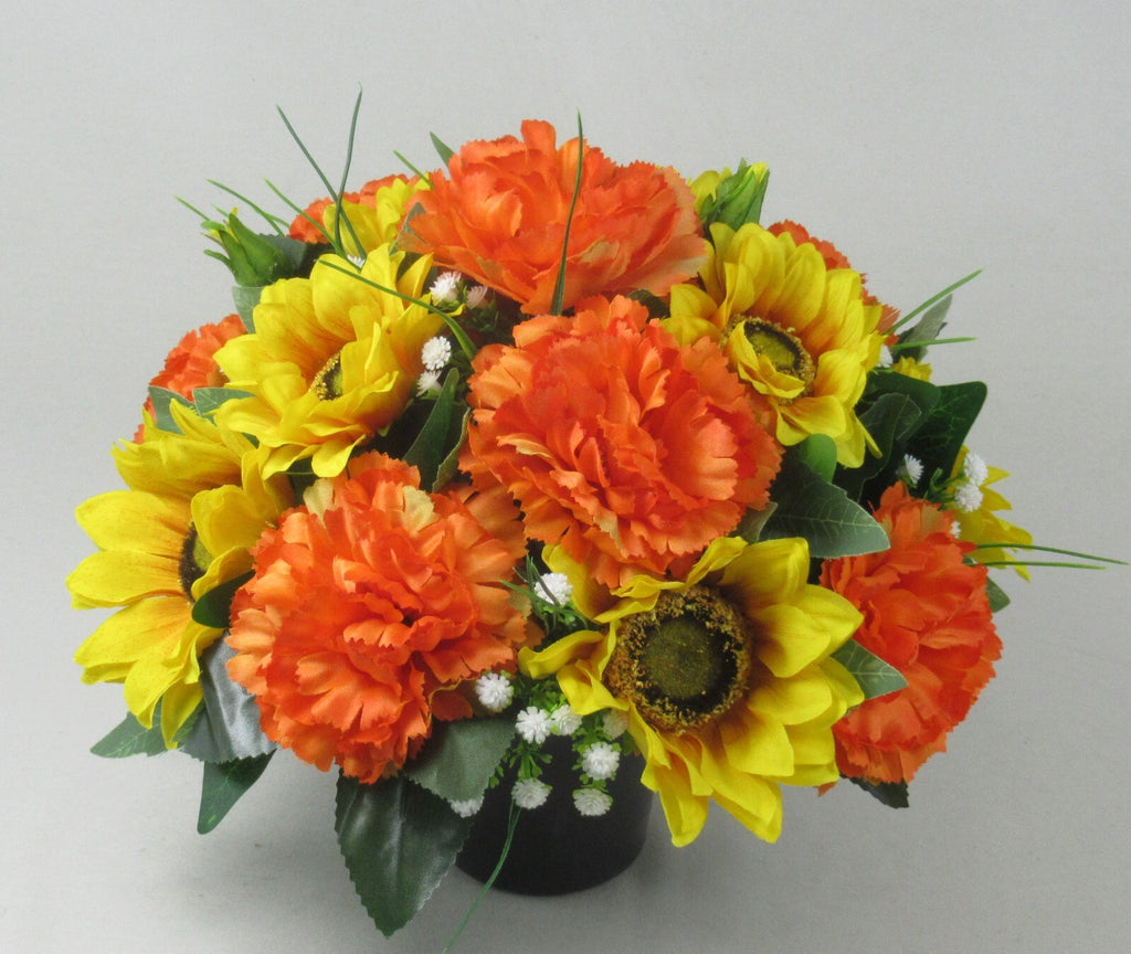 Artificial grave arrangement in dark pot with orange sunflowers, yellow blooms, and white filler
