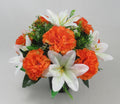 Orange artificial carnations with white lilies in green foliage on black memorial pot
