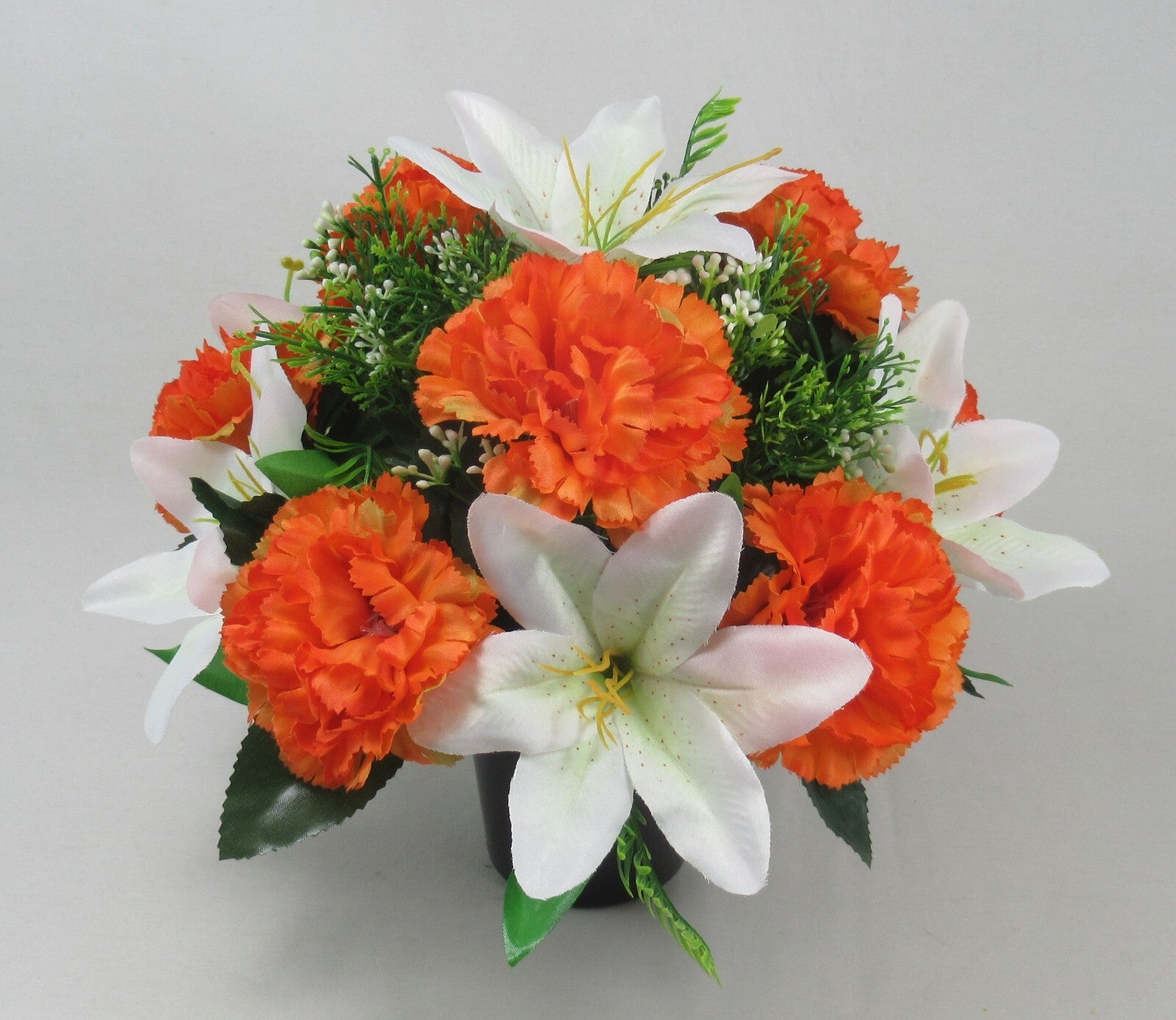 Orange artificial carnations with white lilies in green foliage on black memorial pot