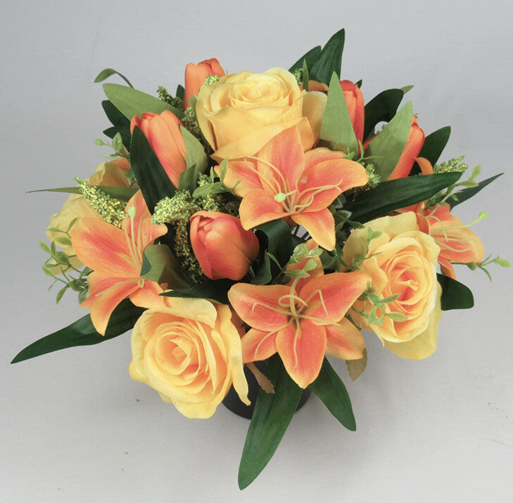 Artificial memorial flowers: yellow roses, orange lily petals, and green leafy stems in a cluster