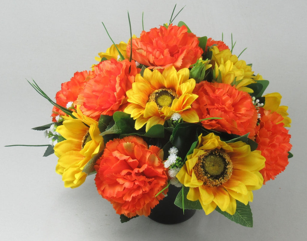 Artificial grave pot arrangement of orange carnations and yellow sunflowers with small white flowers
